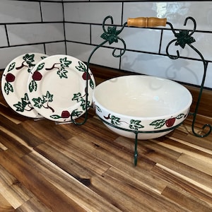 May include: A white ceramic bowl and two plates with a green and red fruit design. The bowl is held by a green metal stand with a wooden handle. The plates are on a clear stand. The items are on a wooden surface.