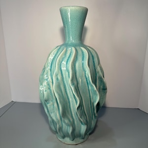May include: A turquoise ceramic vase with a textured, rippled design. The vase has a narrow neck and a bulbous body with vertical wave-like patterns. The glaze has a crackle effect, adding to its unique appearance.
