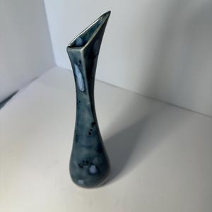 May include: A tall, slender vase with a unique, angled opening. The vase has a teardrop shape and is glazed in shades of blue and gray, with darker spots and lighter streaks. The design is modern and artistic.