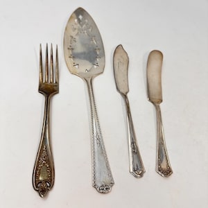 May include: A set of four silver-toned metal utensils including a four-tined fork, a large pointed cake server with decorative cutouts, and two small butter knives. Each piece features intricate embossed floral and geometric handle detailing.