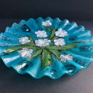 May include: A round, teal-coloured glass plate with a ruffled, wavy edge. The centre features a hand-painted design of white flowers with yellow centres and green leaves. The glass has a textured, speckled appearance with a glossy finish.