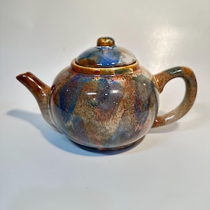 May include: A ceramic teapot with a rounded body and a lid. The teapot features a speckled glaze in shades of blue, brown, and orange. The handle and spout are a solid brown color. The lid has a gold-colored knob. The text "SP-BEAN PORT-JOL" is visible on the teapot.