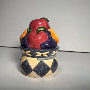 May include: A ceramic jar with a lid shaped like a basket of fruit. The jar has a cream-colored base with navy blue diamond patterns. The lid features red apples, purple grapes, and yellow pears.