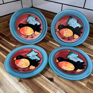 May include: Four ceramic plates with a teal rim and a red center. Each plate features a black bird with an orange breast and a green tree design. The plates are arranged on a wooden surface.