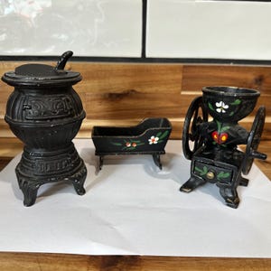 May include: A set of three miniature cast iron decorative items: a stove, a cradle, and a coffee grinder. The stove and grinder are black with ornate details. The cradle is black with painted floral accents.