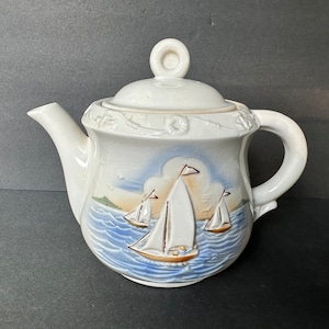 May include: White ceramic teapot with a nautical design. The teapot features a lid with a round handle and a spout. The body of the teapot is decorated with a scene of sailboats on the sea, with blue and brown accents.
