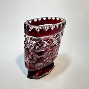 May include: A ruby red, cut crystal vase with a unique oval shape. The vase features intricate geometric and floral patterns, with a scalloped rim. The base is a solid, faceted shape. The vase is approximately 6 inches tall.