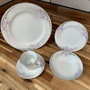 May include: A white porcelain dinnerware set featuring a floral design with pink and purple flowers and gray accents. The set includes a large dinner plate, a smaller plate, a bowl, and a teacup with a saucer.