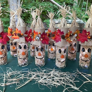 May include: A collection of clear glass bottles filled with straw, each featuring a hand-painted scarecrow face. The neck of each bottle is wrapped in burlap and decorated with artificial autumn leaves, pinecones, and small orange berries.