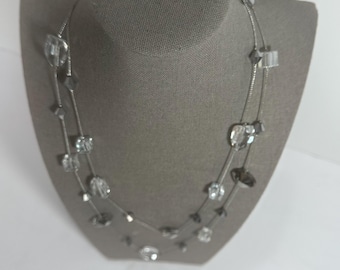 May include: A two-strand necklace with clear and gray beads. The necklace is displayed on a gray mannequin. The beads are various shapes, including squares, diamonds, and rounded shapes. The necklace is silver in color.