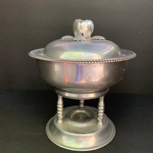 以下が含まれることがあります： 蓋とスタンドが付いた銀製のサービングディッシュ。蓋には装飾的な花の模様が施されています。ディッシュは3本の脚のスタンドの上に置かれています。