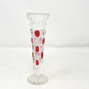 May include: A clear glass bud vase featuring a flared rim and a tapered body with a diamond-cut pattern. The exterior is decorated with vertical rows of inset, smooth, circular red glass cabochons. The base is circular with a starburst design.