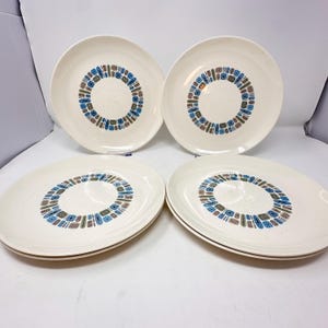 May include: A set of four round, off-white ceramic dinner plates. Each plate features a central circular pattern composed of small, repeating geometric shapes in shades of blue, brown, and black, arranged in a ring around the center of the plate.