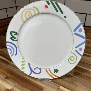 May include: White ceramic plate with a colourful abstract design around the rim. The design includes green, blue, orange, yellow, and purple shapes. The plate has a silver trim and is displayed on a wooden surface.