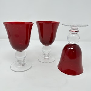 May include: Three wine glasses with a clear, round base and stem. The bowl of the glasses is a deep red color. One glass is upside down. The glasses are on a white surface.