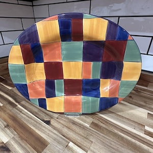 May include: A large, oval-shaped ceramic platter with a colorful patchwork design. The platter features squares of orange, yellow, green, blue, and burgundy. The rim is a dark blue. The platter is on a wooden surface.