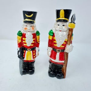 May include: Two ceramic nutcracker figurines wearing black hats, red jackets with yellow accents, and black boots. One figure holds a black sword, while the other holds a tall wooden spear. Both have white beards and painted facial features.