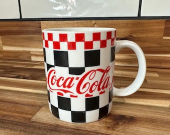May include: White ceramic Coca-Cola mug with a red and black checkered pattern. The Coca-Cola logo is in red script across the front. The mug has a white handle and is on a wooden surface.