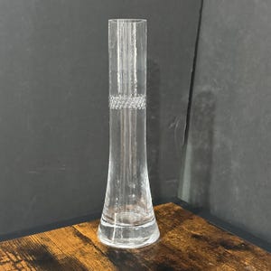 May include: A clear glass vase with a cylindrical shape and a flared base. The vase features a textured surface and a decorative band of small, raised dots around the middle. The vase is approximately 10 inches tall.