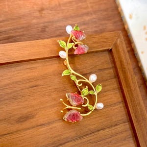 Puede incluir: Un pendiente de oreja dorado con un diseño floral. Presenta flores rojas y rosas, hojas verdes y detalles de perlas blancas. La joya está colocada sobre un fondo de madera.