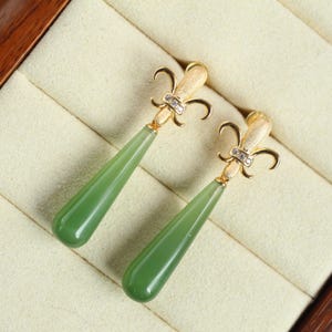 Può includere: Un paio di orecchini pendenti color oro con un design a giglio. Ogni orecchino presenta una pietra a forma di goccia verde chiaro. Gli orecchini sono esposti in un portagioie color crema.