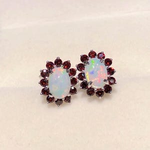 May include: A pair of oval opal stud earrings. Each earring features a central, iridescent opal stone surrounded by a halo of small, dark red gemstones. The earrings are set in a silver-colored metal.