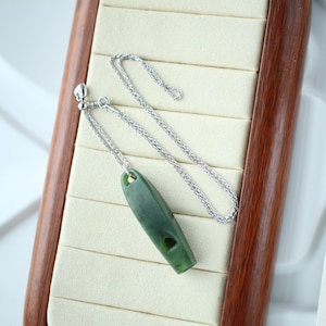 Op de afbeelding: Een ketting met een donkergroene jade hanger en een zilveren ketting. De hanger heeft een langwerpige ovale vorm met een klein gaatje. De ketting wordt gepresenteerd op een crèmekleurig oppervlak in een houten frame.