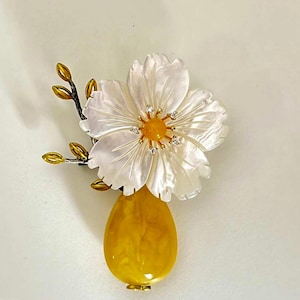 May include: A floral brooch featuring a white flower with a mother-of-pearl appearance, accented with small diamonds and a yellow center. The flower sits atop a yellow, teardrop-shaped base, with gold-colored accents and a branch with gold buds.