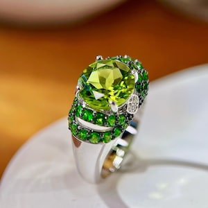 May include: A silver ring featuring a large, oval-cut, light green gemstone as the centerpiece. The ring is further adorned with smaller, round, dark green gemstones and a cluster of small, clear stones, creating a luxurious and eye-catching design.