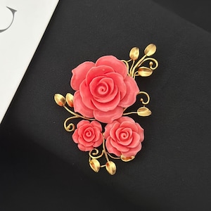 May include: A coral-colored rose brooch with three roses and gold-colored leaves and stems. The brooch is set against a black background. The roses are in full bloom, with detailed petals.