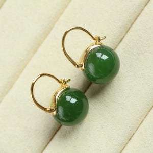 May include: A pair of gold-coloured hoop earrings featuring polished, dark green gemstone beads. The earrings are set against a cream-coloured background, highlighting the jewellery's design and colour.