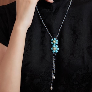 Puede incluir: Un collar largo con colgante de flor turquesa, con cadena dorada y detalles de perlas. El collar está hecho de cuentas de vidrio azul y presenta dos flores turquesas con centros de perlas.