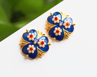 May include: A pair of gold-tone earrings with blue enamel and red and white pearl accents. The earrings are shaped like a four-leaf clover.