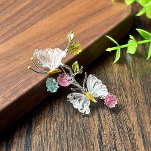 Può includere: Spilla color argento con design a farfalla e fiori. La spilla presenta dettagli trasparenti, rosa, gialli e verdi. La farfalla ha ali trasparenti e dettagli gialli. I fiori sono rosa, gialli e blu. La spilla è su una superficie di legno.