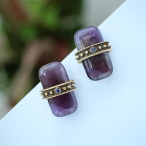 May include: Two amethyst-colored stone earrings with gold-colored bands. Each earring has a rectangular shape with a gold band featuring small, round accents and a small blue stone in the center. The earrings are set against a white background.