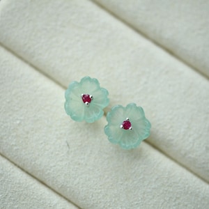 May include: A pair of flower-shaped stud earrings. Each earring features a light blue, translucent floral design with a small, round, red gemstone center. The earrings are set against a white, textured background.