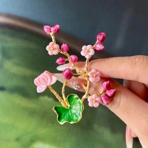Peut inclure: Broche florale avec une branche dorée ornée de fleurs roses et fuchsia, de boutons et d'une feuille verte. La broche est munie d'une épingle pour la fixer aux vêtements ou accessoires. Design délicat et détaillé.