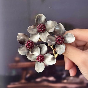 May include: A silver-toned floral brooch featuring five flower blossoms with dark red centers. The brooch has a gold-colored stem and is held against a blurred background.