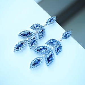 May include: A pair of elegant blue leaf-shaped earrings. Each earring features a silver-toned metal base adorned with numerous small, round blue crystals and larger, marquise-cut blue gemstones, creating a sparkling, nature-inspired design.