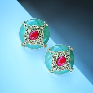 May include: A pair of round earrings with a teal-green base and a central red oval. The earrings feature a gold-colored filigree design around the red center and the outer edge. The earrings are set against a gradient background.