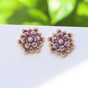 May include: A pair of gold earrings with a flower design. The earrings are set with purple and white gemstones.