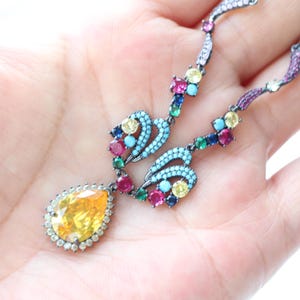 May include: A colorful necklace with a large yellow teardrop-shaped pendant. The necklace features a variety of gemstones in shades of yellow, pink, blue, and green. The metal is dark, and the design is intricate and ornate.