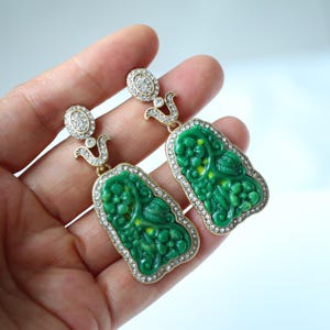 Puede incluir: Un par de pendientes verdes con un diseño floral. Los pendientes tienen un marco dorado y están incrustados con pequeños cristales transparentes. Los pendientes son rectangulares con una parte superior curvada y un gancho decorativo dorado.