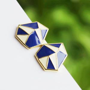 May include: A pair of geometric stud earrings with a white and blue enamel design. The earrings are gold-toned and have a faceted shape.