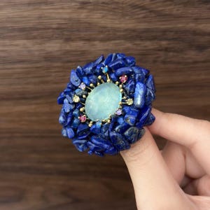 May include: A unique ring featuring a large, oval, pale blue gemstone at the center, surrounded by a cluster of deep blue lapis lazuli stones. Small, colorful gemstones and gold accents add detail to the design.