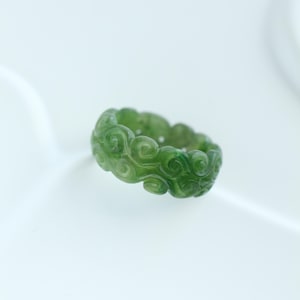 May include: A green jade ring with a swirling cloud design. The ring is carved with intricate details, showcasing a continuous pattern of flowing curves. The ring is set against a white background.