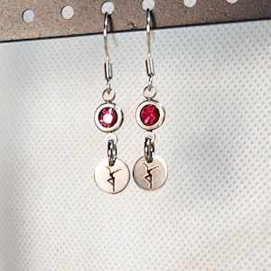 May include: Silver dangle earrings with a red gemstone and a small, round charm featuring a dancer silhouette. The earrings hang from a silver hook.