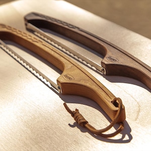 May include: Two wooden bread knives with serrated blades and leather lanyards. One knife is light-colored wood, the other is dark. The handles have a curved design and the brand name "DANO" is visible on the handles. The knives are on a light wood surface.