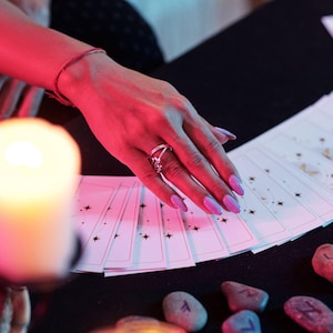 May include: A hand with purple nails and a silver ring touches a deck of tarot cards. The cards are white with black star designs. A lit candle and several rune stones are also visible.