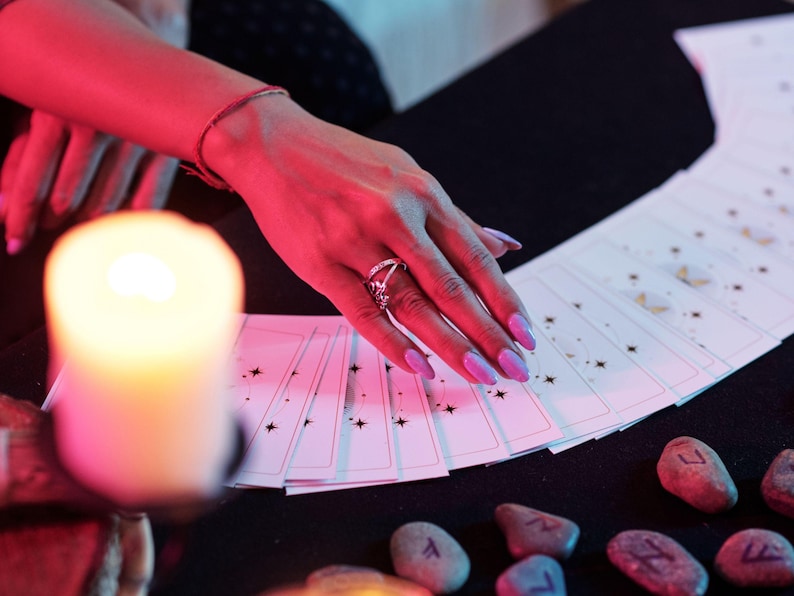 May include: A hand with purple nails and a silver ring touches a deck of tarot cards. The cards are white with black star designs. A lit candle and several rune stones are also visible.